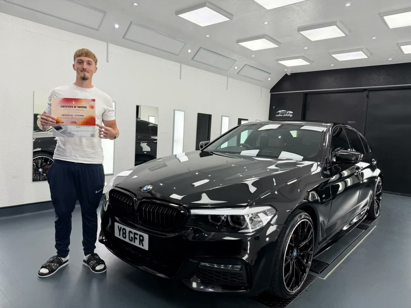 Grandeur Autocare detailer holding a certificate of training in 3-stage paint correction and ceramic coating, next to a black BMW 5 Series